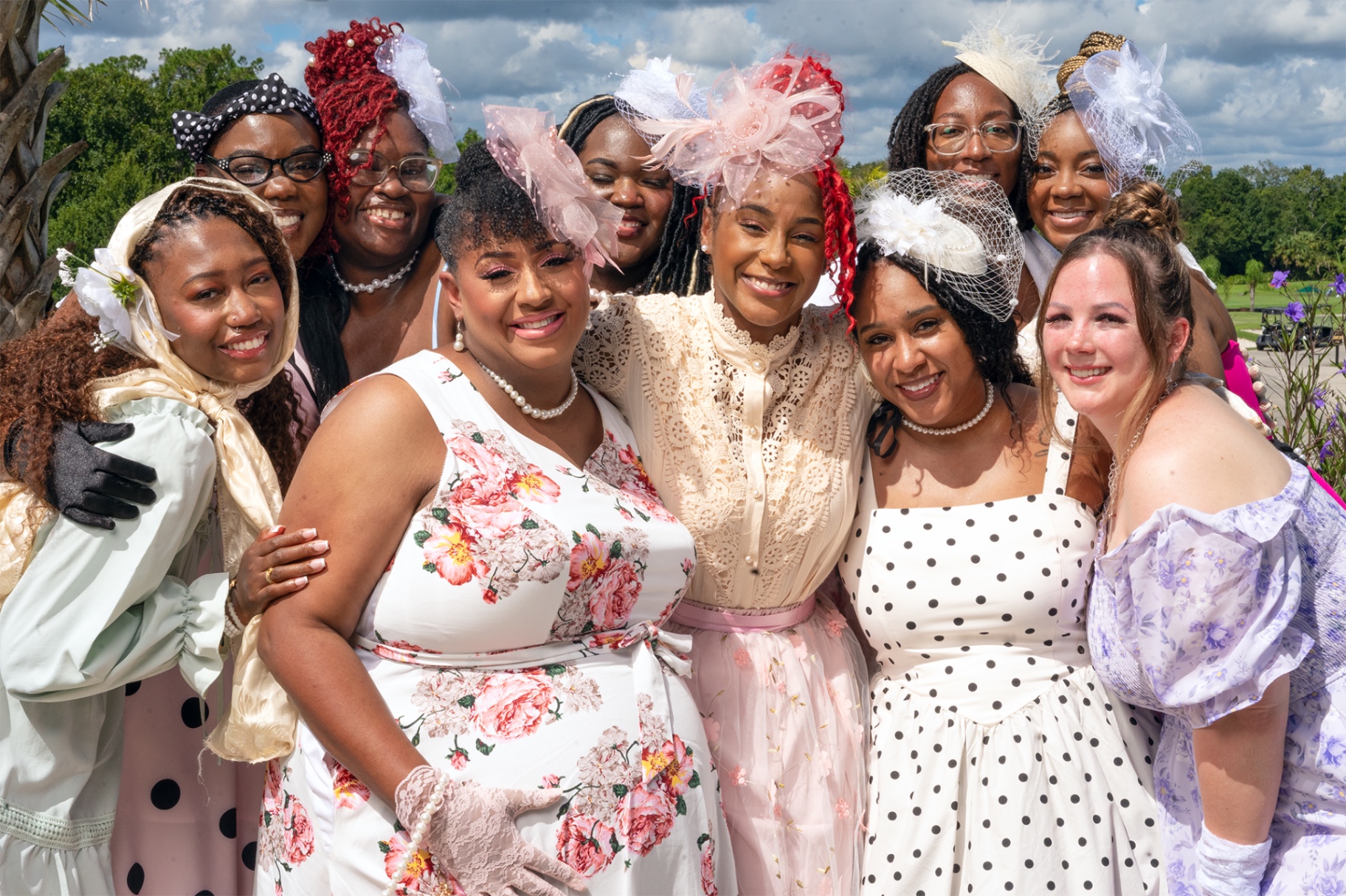The Bride Tribe women in tea-party dresses and fascinators smiling together in a sunny garden